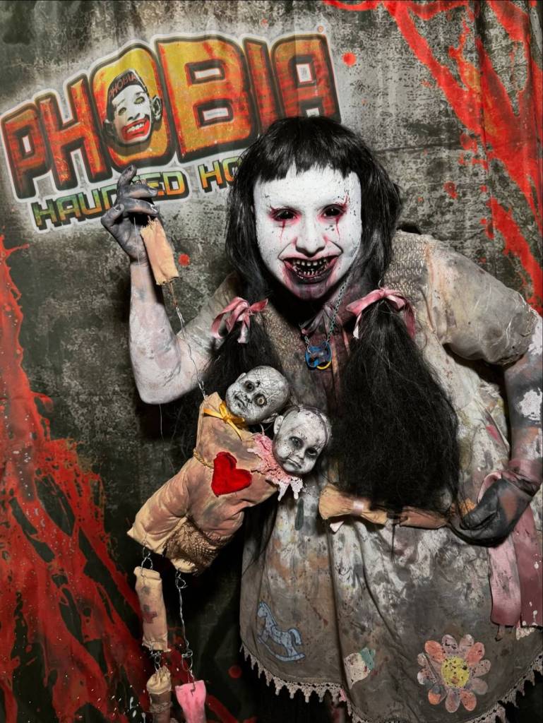 La imagen muestra una espeluznante figura de muñeco posando delante de un cartel de Phobia Haunted Houses. 