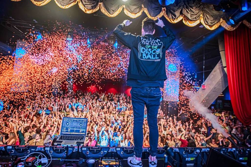 ein DJ steht mit erhobenen Armen auf der Bühne von Gimme Gimme Disco, Konfetti wird ins Publikum geschossen und die Leute auf der Tanzfläche jubeln mit ausgestreckten Armen