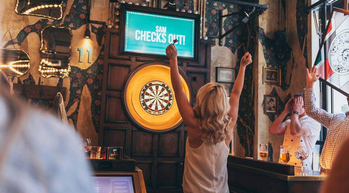 A woman celebrates after throwing a dart at a target. A screen above the oche reads "Sam checks out!"