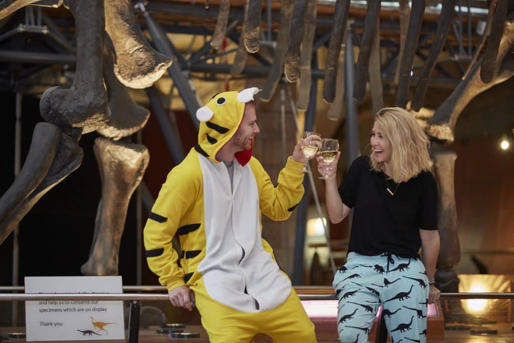 two adults, one in a tiger onesie, clinking glasses of wine as they enjoy the dino snores event
