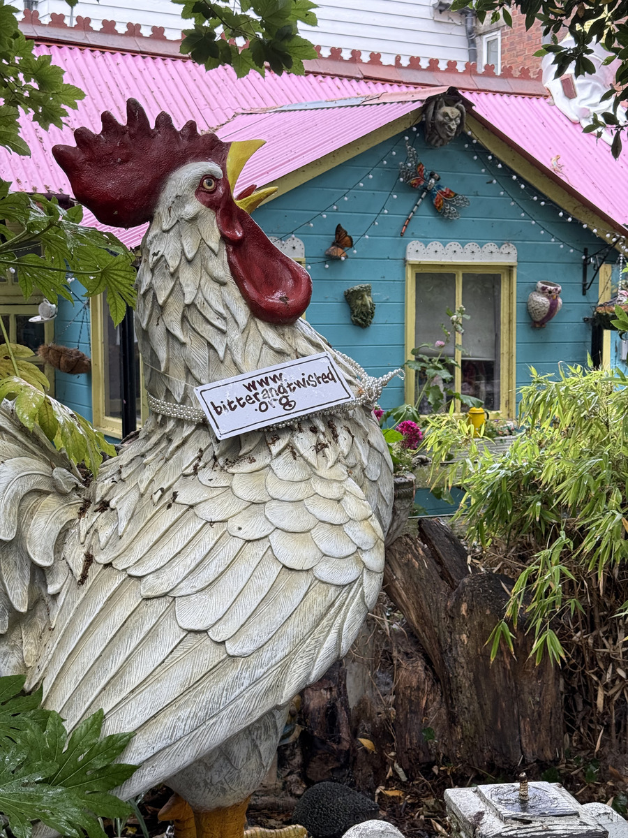 an enormous iron chicken statue in front of a green building with a pink roof