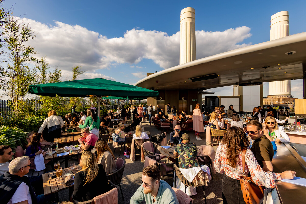 a rooftop bar in london with people relaxing in the sunshine and battersea power stations iconic chimneys visible in the background