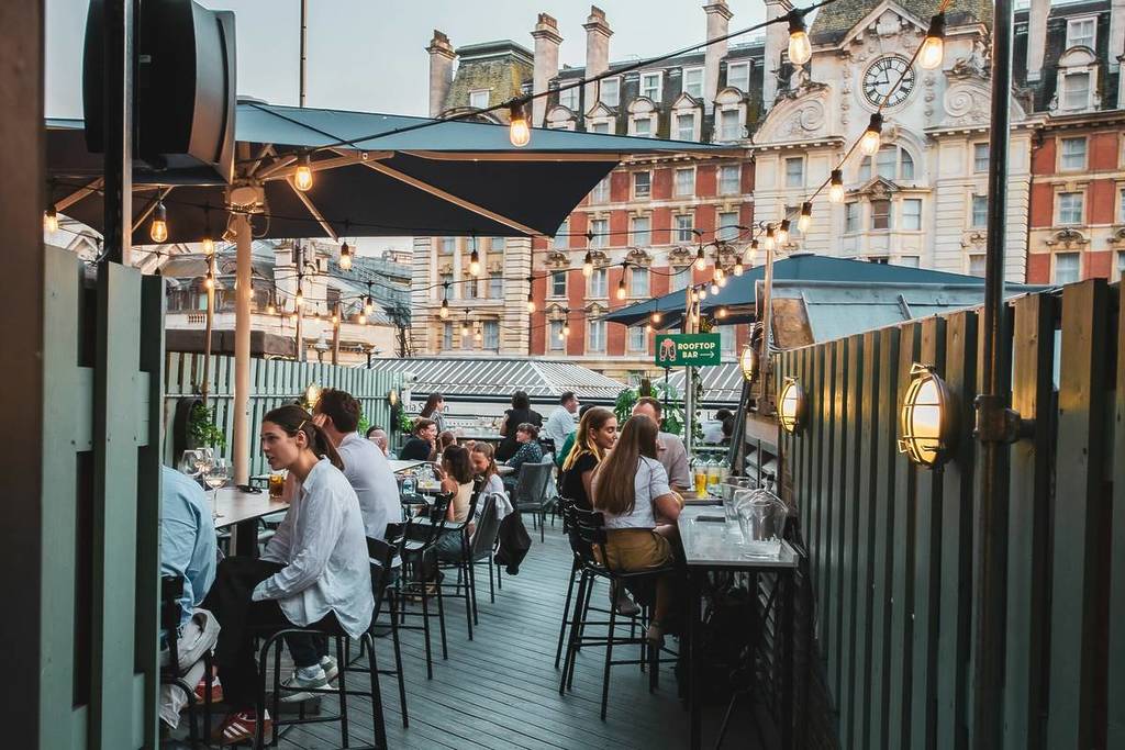 an outdoor terrace filled with people at tables. a sign in the back reads 'rooftop bar' with an arrow pointing to the right.