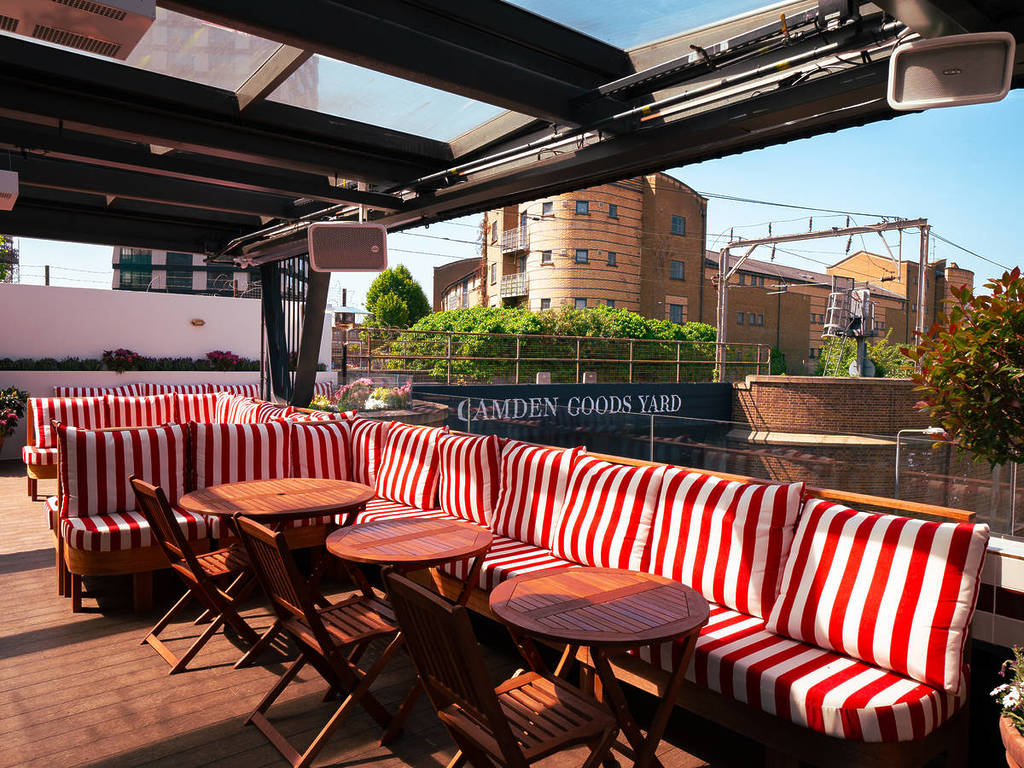 A sunny rooftop with red and white striped booth seating, wooden tables, and a railway in the background.