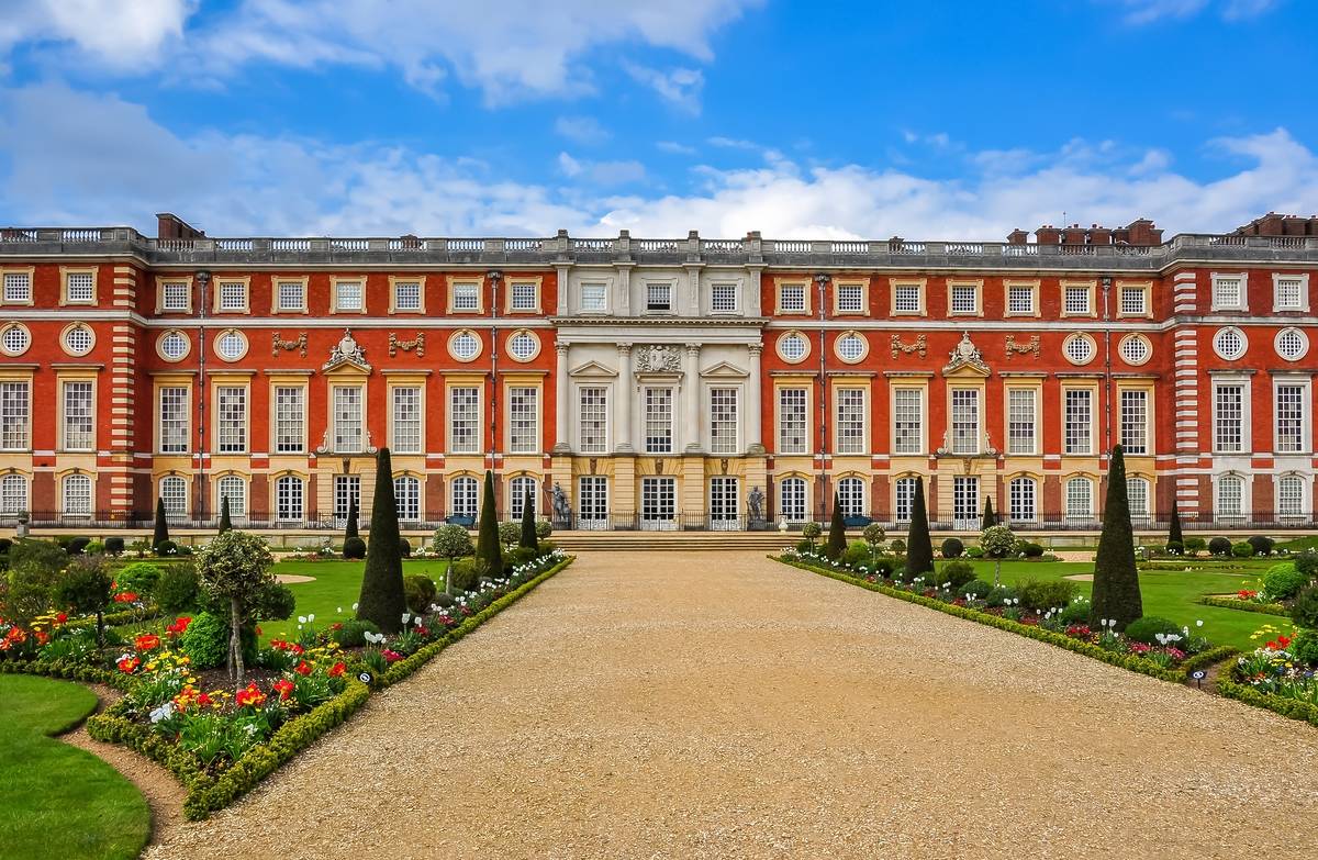 the red exterior of hampton court palace on a sunny blue sky day, with gardens visible along the path leading to the entrance