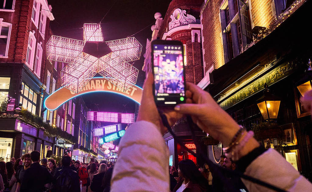 qualcuno che di notte scatta una foto con il cellulare alle luci natalizie al neon dalle forme spigolose di Carnaby Street