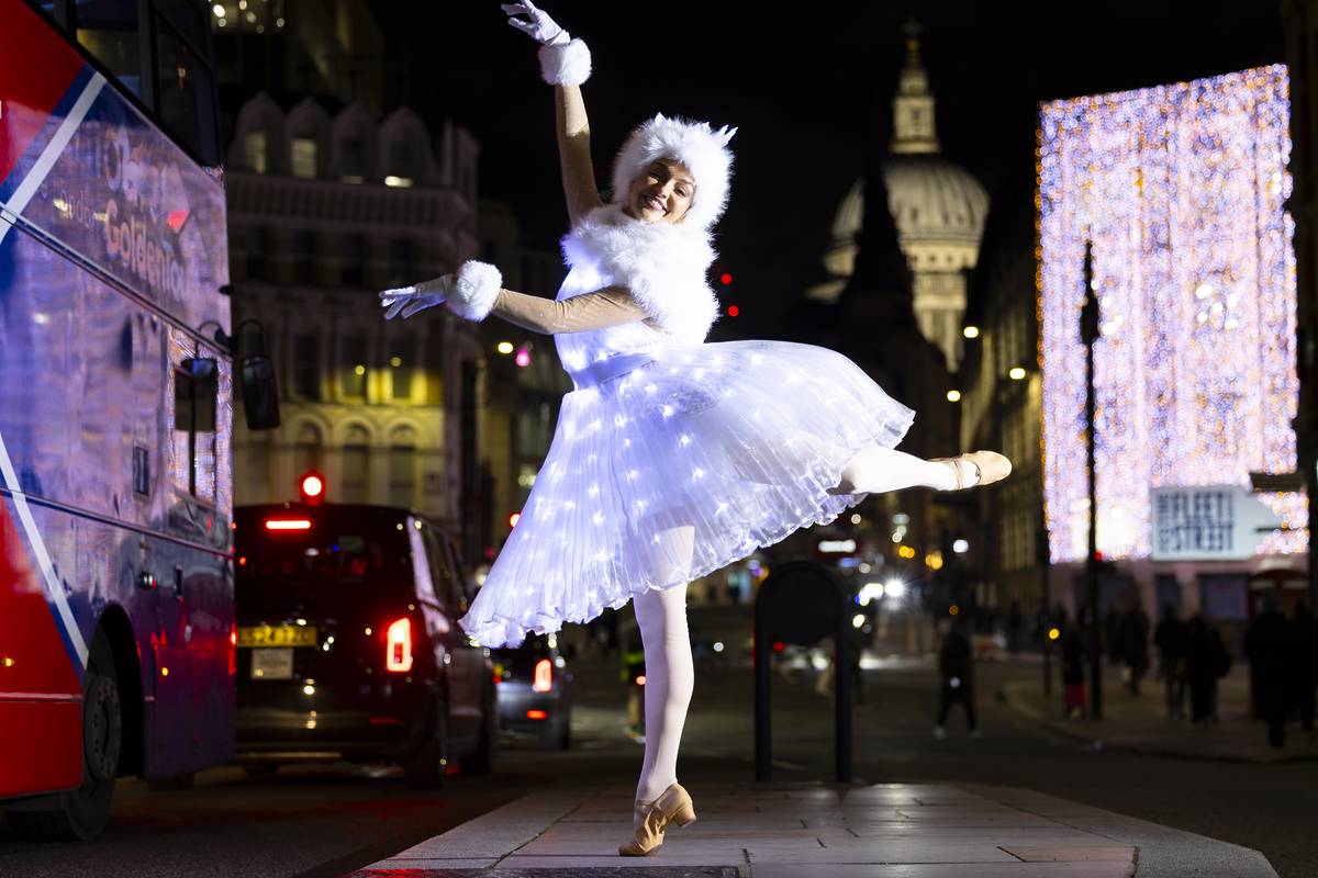 a ballerina striking a pose in a wintry outfit in front of the fleet street quarter christmas lights