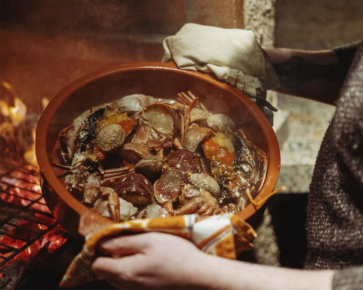 an over the shoulder shot of a cook holding a large earthenware pot full of cooking shellfish