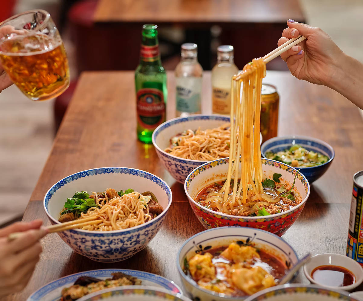a spread of bowls of fiery noodles, wontons, and drinks on a table, with two people enjoying the food and drink