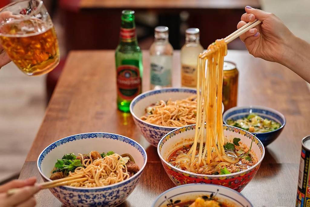 eine Reihe von Schüsseln mit feurigen Nudeln, Wontons und Getränken auf einem Tisch, auf dem zwei Personen das Essen und die Getränke genießen
