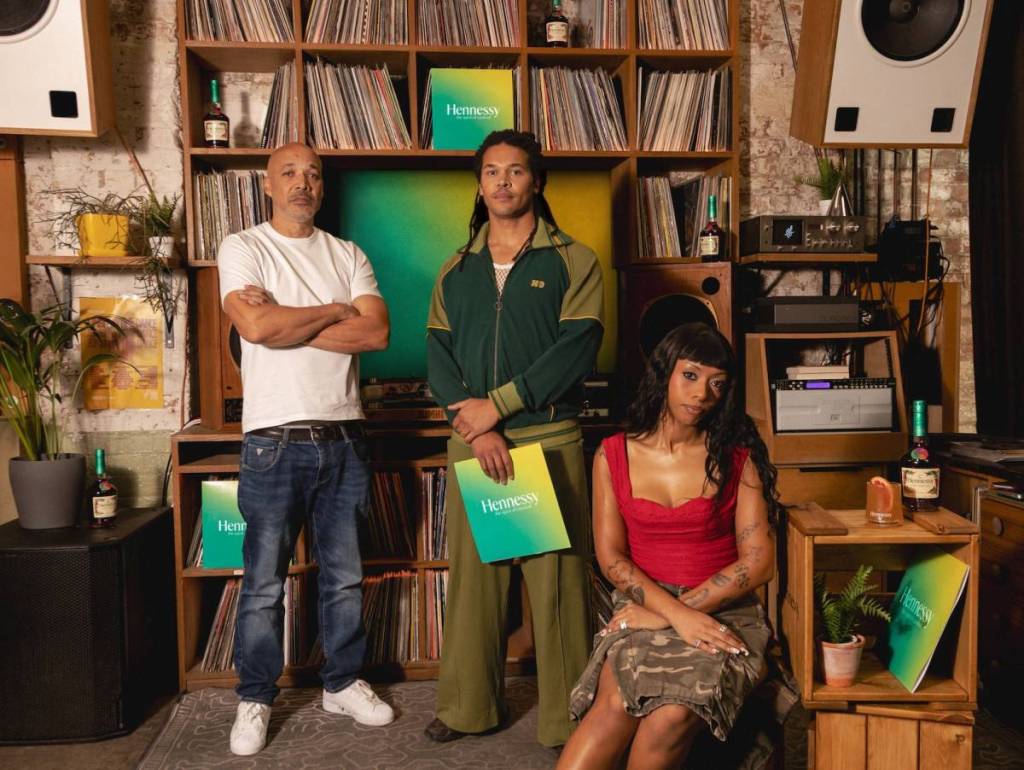 três artistas posam em frente a uma prateleira de discos; o do meio segura um disco azul e amarelo com a inscrição «Hennessey»
