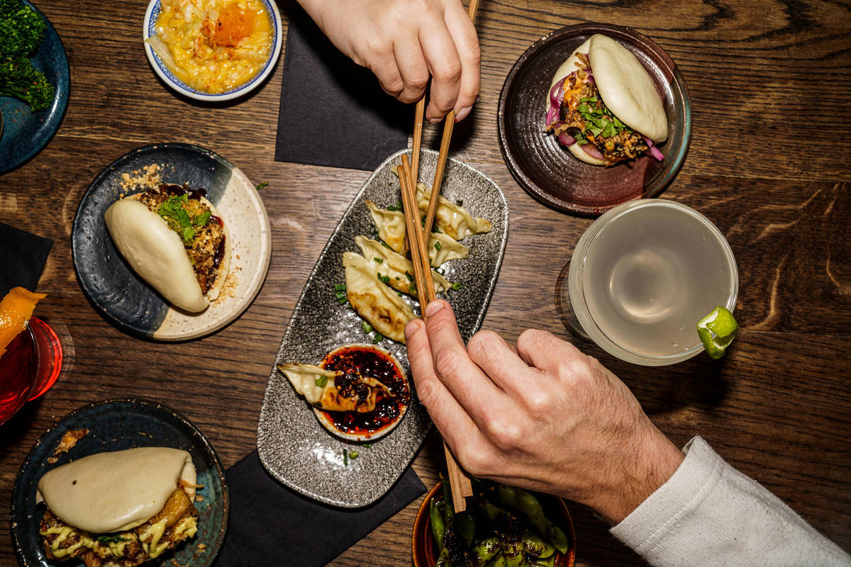 two hands wielding chopsticks, fighting to grab a gyoza from a platter, which is surrounded by plates of bao and cocktails