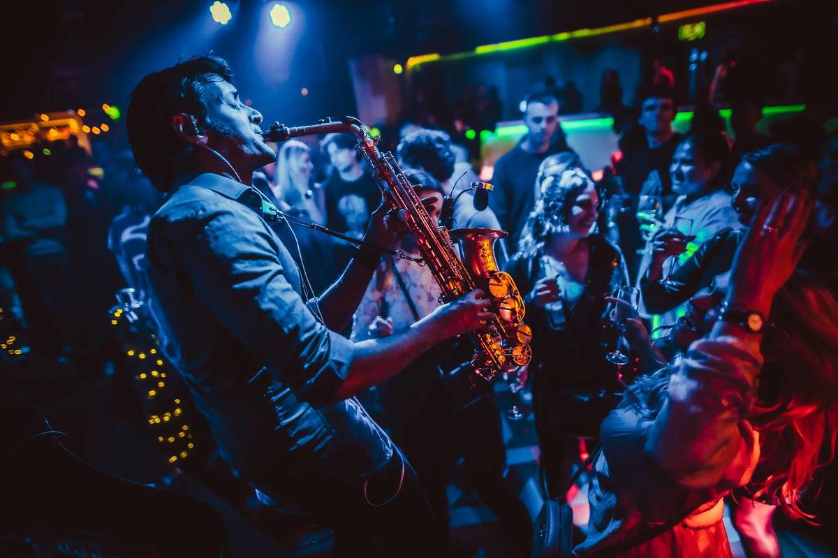 people dancing in a low-lit, blue and red hued, bar with drinks in hand around someone playing a saxophone