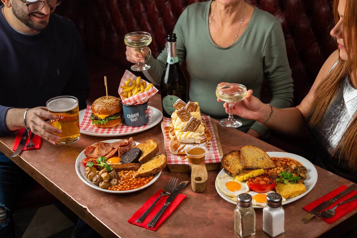 people holding glasses of prosecco and pints of beer around a table spread with breakfast delights like pancakes and full english breakfasts