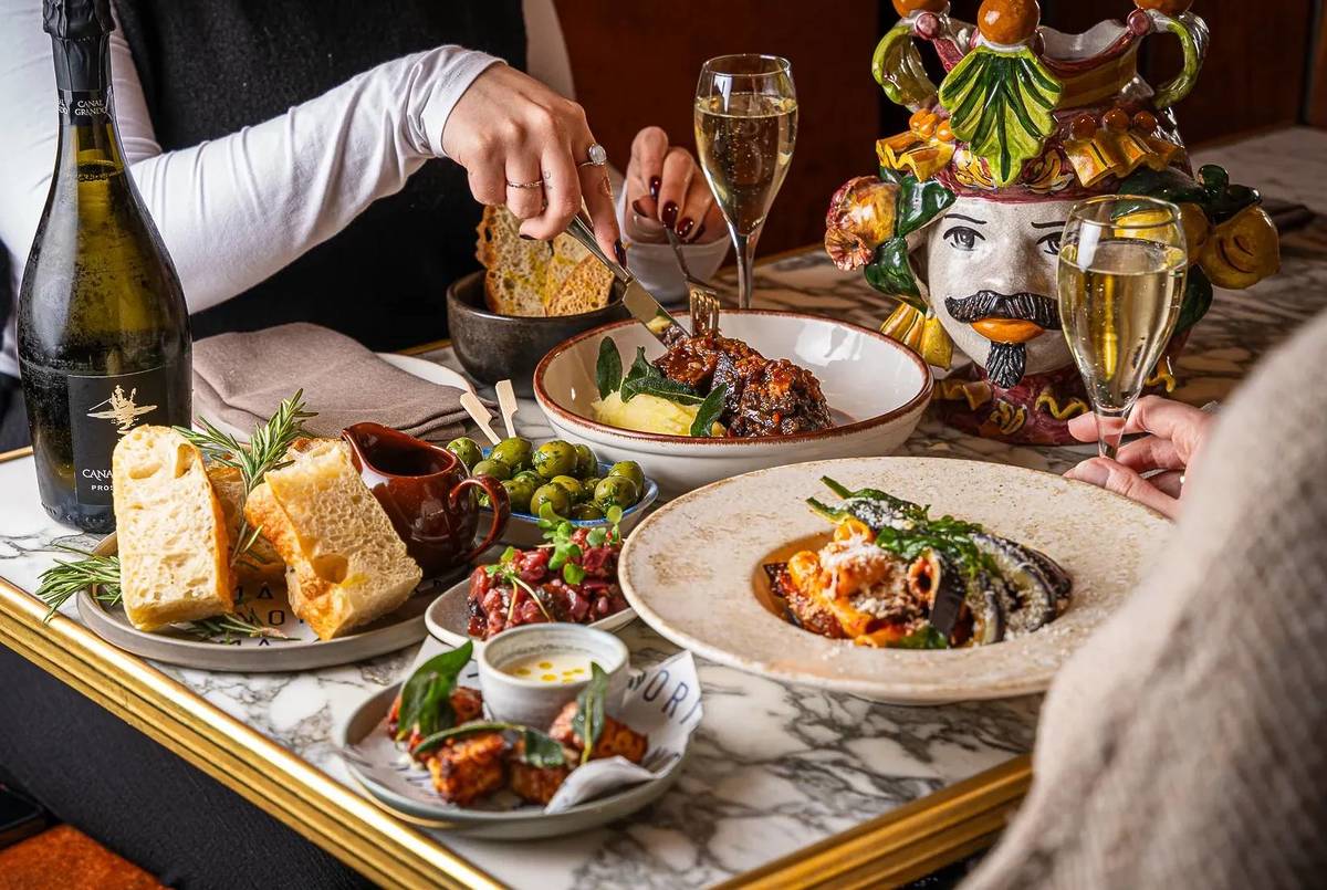 two people tucking into hearty italian dishes from a table covered in food - with platters of focaccia and olives and sides and glasses of prosecco