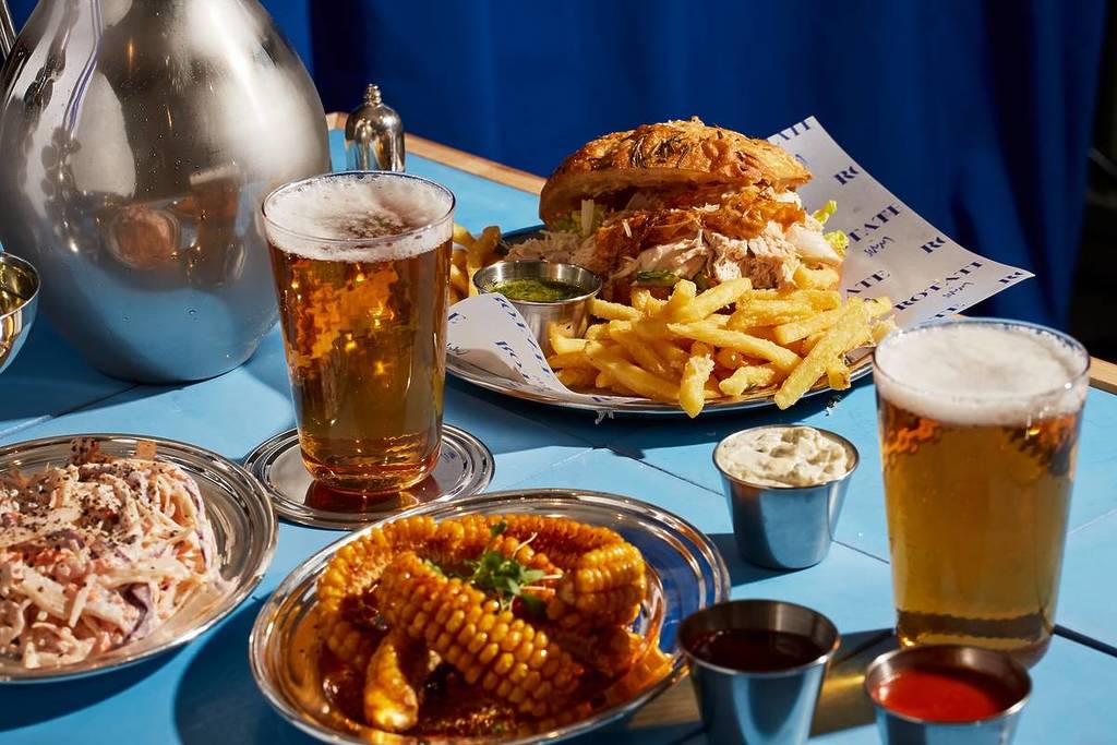 une sélection de plats du restaurant Jimmy Garcia Rotate by Jimmy - notamment du maïs en épi, un burger au poulet rôti, des frites et de la bière dorée 
