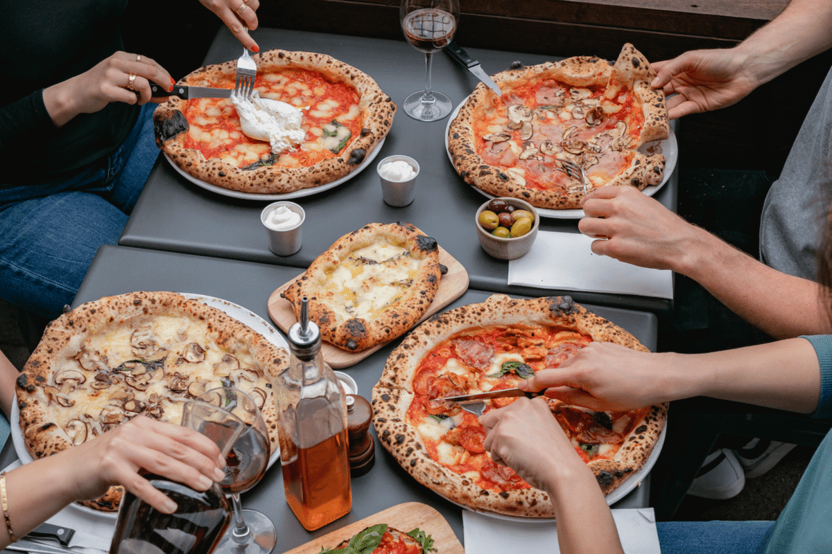 Four pairs of hands cutting into and picking up slices from a delicious spread of pizzas for the bottomless brunch at Crust Bros.
