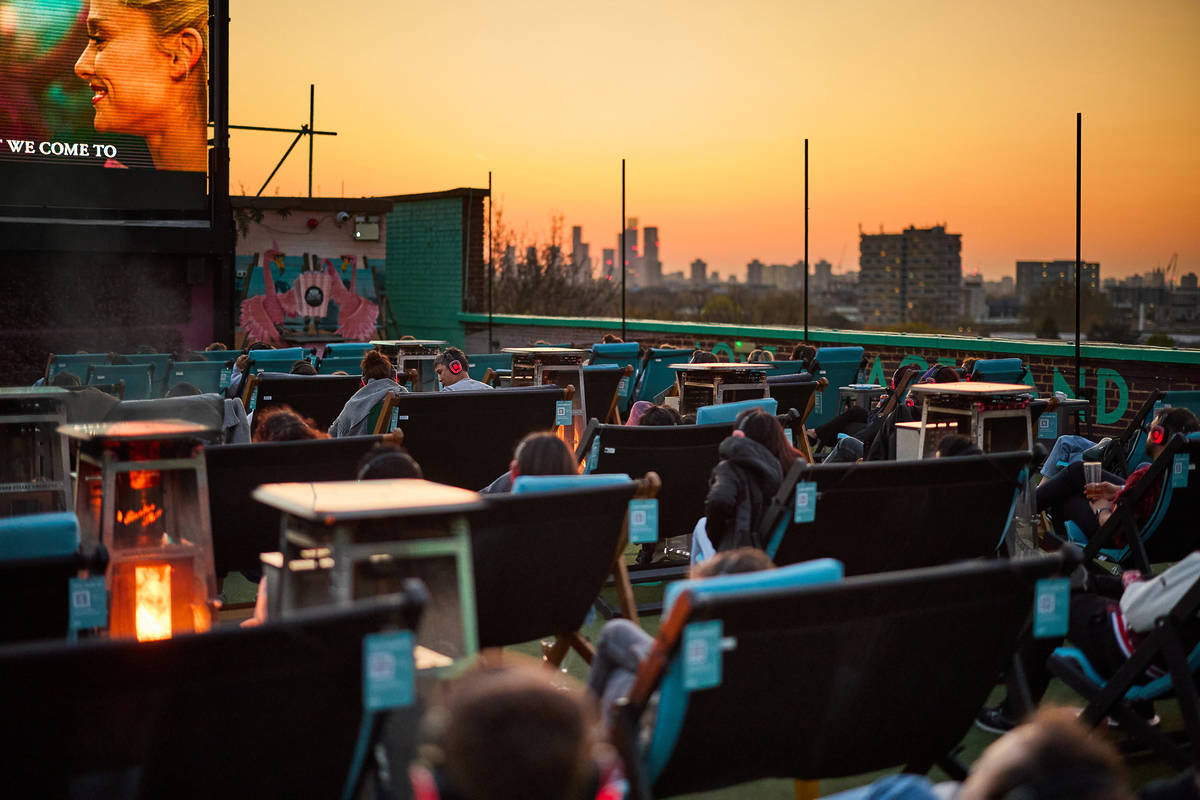 people in sun comfy chairs on a rooftop watching a movie on a large screen with a view of the london skyline in the background