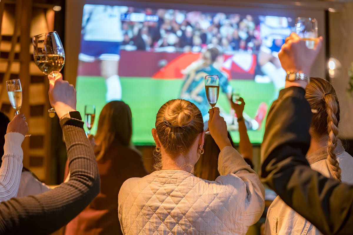 people holding up their glasses in celebration at a large screen showing a football match