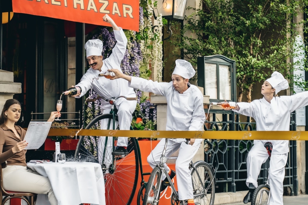 An image of the tour de rose interactive experience at Balthazar, where three chefs are trying to pour wine while seated on bicycles