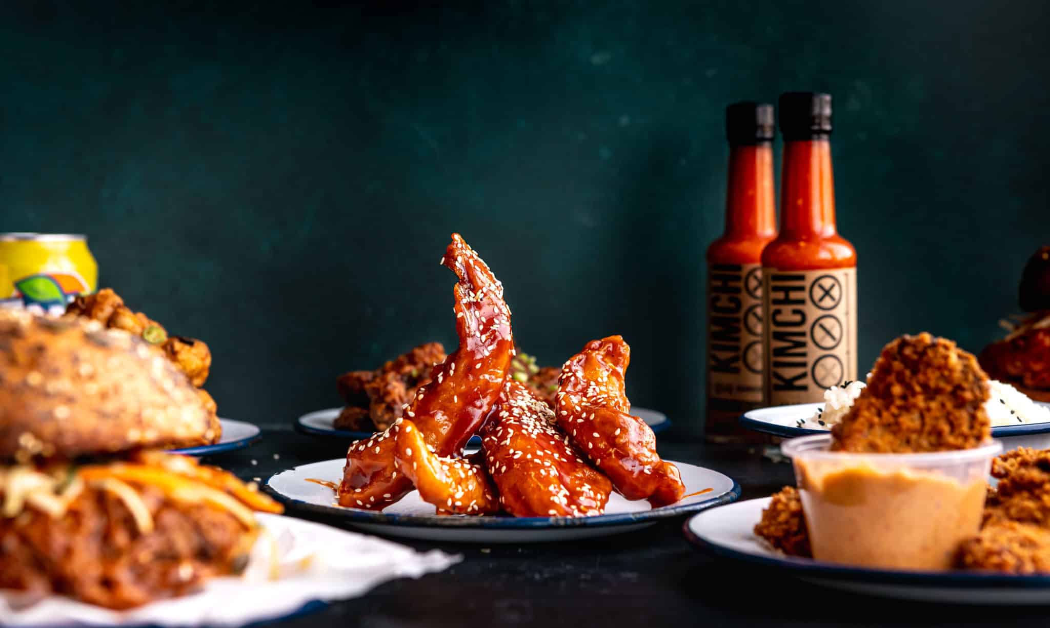 a burger next to fried chicken tenders with two bottles of hot sauce visible in the background