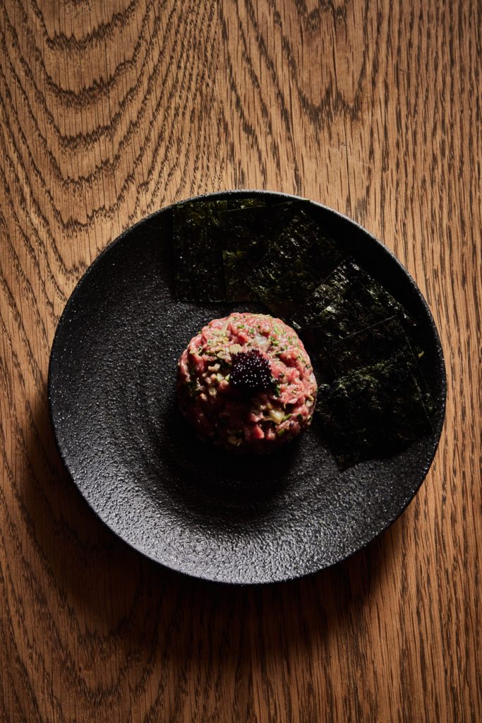 a black plate of beef tartare and nori sheets, atop a wooden table