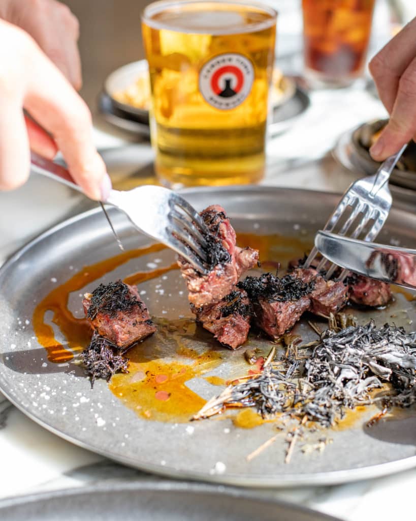 forks and knives cutting into a steak, with a beer in the background, forming part of the beer and steak pairing at the black cow