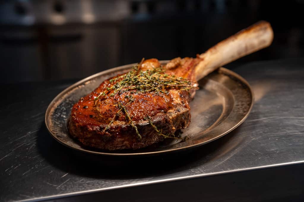 a beautifully cooked steak resting on a broiler plate, a bone sticks out of the back of teh massive cut, which is available on Tomahawk Thursday