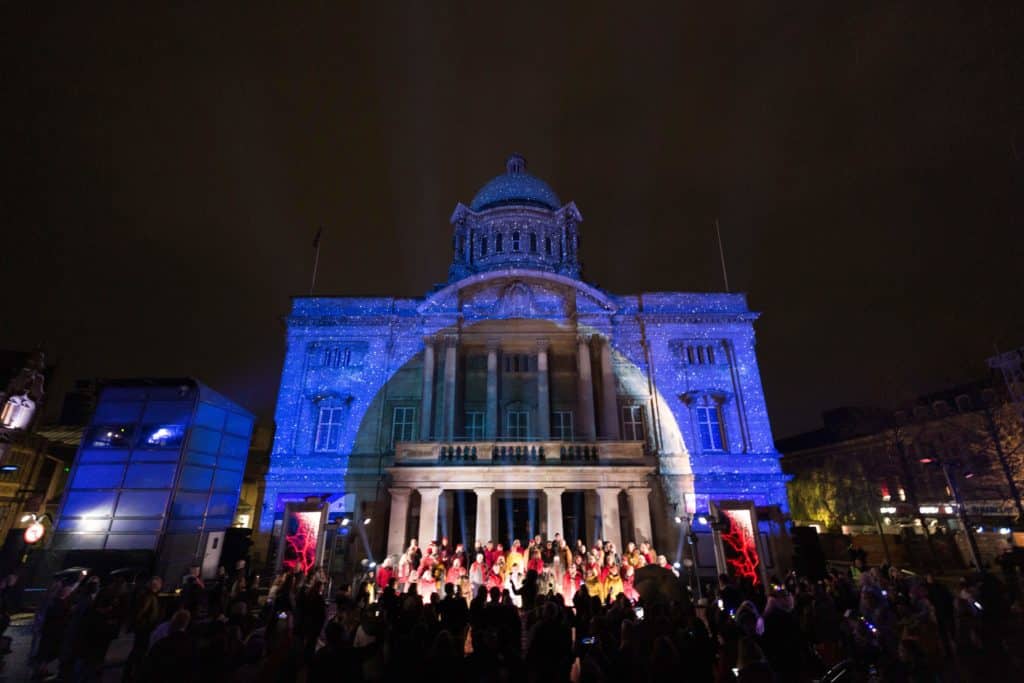 the about us lightshow projected onto a building in hull, with the choir spotlighted by a bright white spherical projection over them