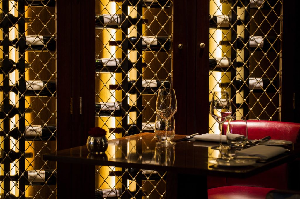 a wall of wine at The Game Bird, backlit by soft yellow lighting, a table is set up in front with wine glasses atop