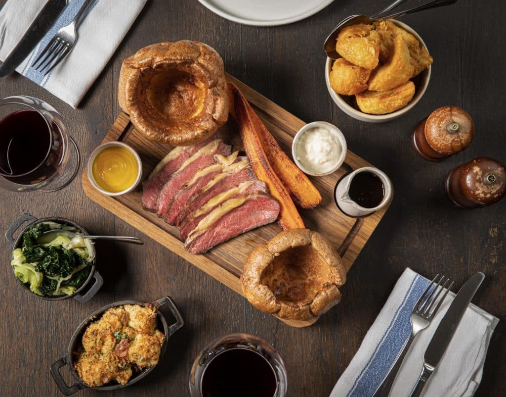 the sunday roast at pig and butcher, showing a wooden board topped with roast beef, yorkshire puddings, carrot, and sauces, with plates and bowls of sides scattered around it