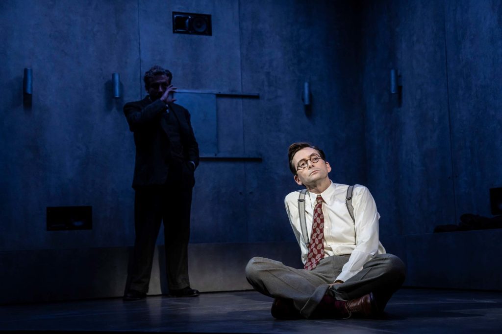 David Tennant on stage, with a shadowy figure behind him, clouded in black and blue lighting