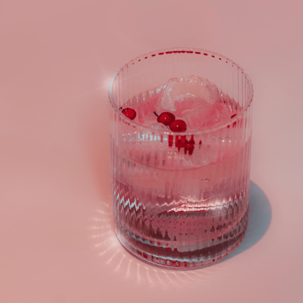 a pink hued cocktail in a rocks glass, with pink peppercorns, on a pink background