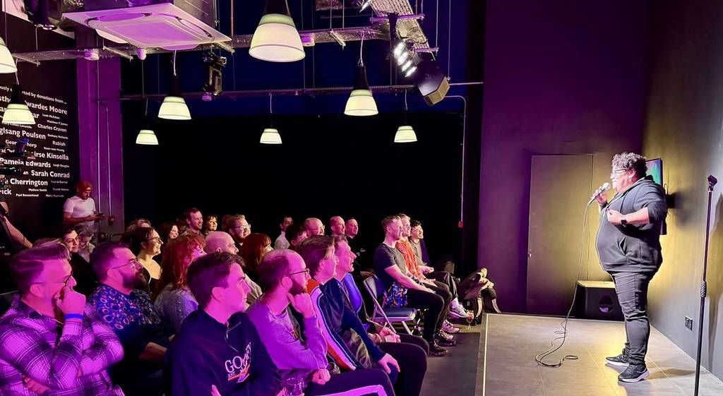 A comedian performing on a stage in front of a seated crowd at a queer comedy venue.