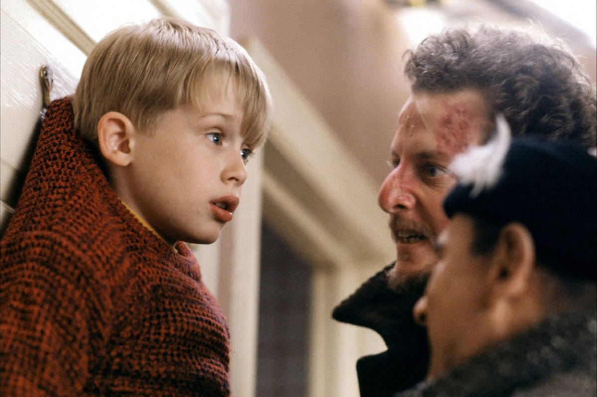 a still from the 'home alone' film showing a young boy dangling from a door while two dirty looking thieves interrogate him