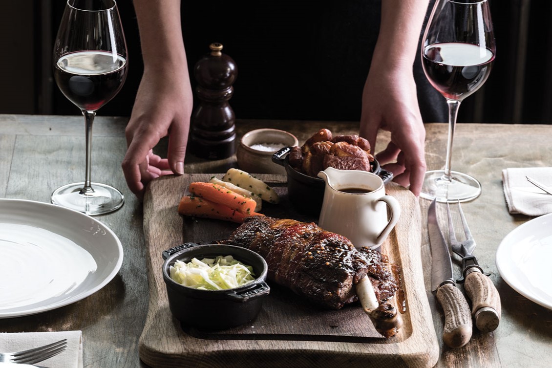 someone setting down a heaving platter containing a beef roast and veg sides, between glasses of wine and plates