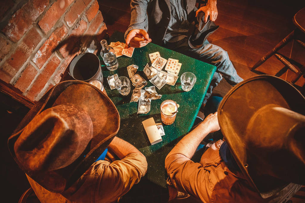 un groupe de personnes vêtues de tenues du Far West/de cow-boys rassemblées autour d’une table où se déroule une partie de cartes