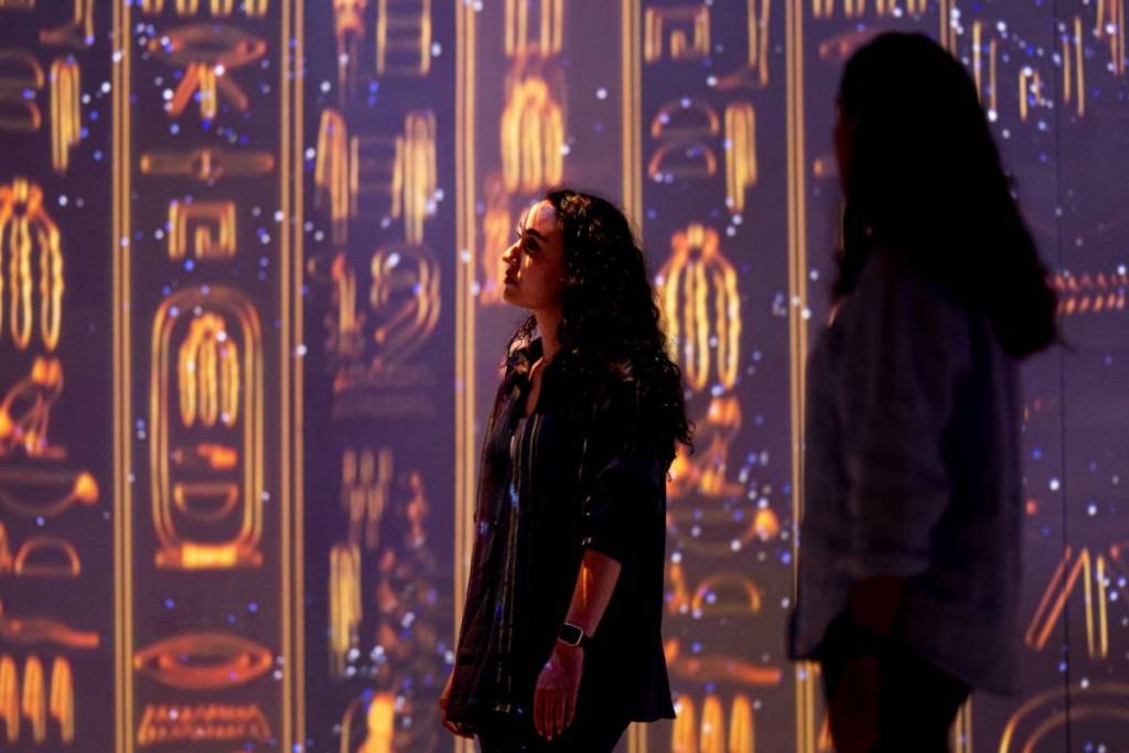 two women look at projections of golden hieroglyphic on the walls at the Timewalk Exhibition