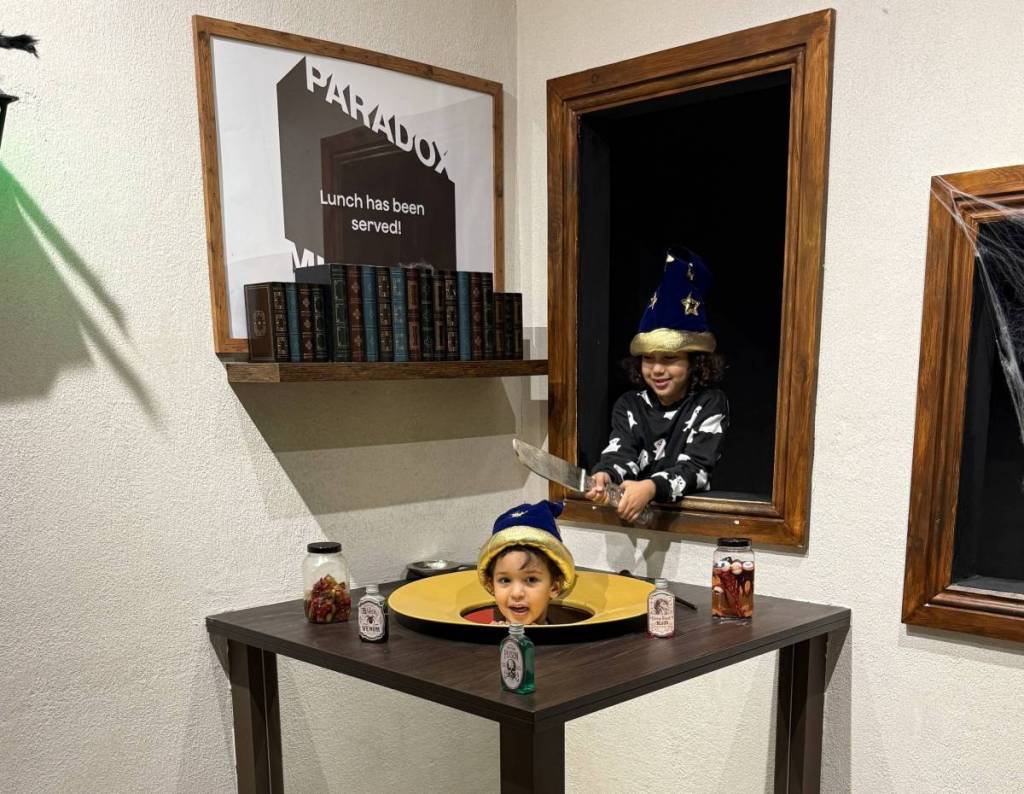 two children in ghost onesies laugh as they pose in an optical illusion where it appears like there is a head sitting on a plate at Paradox Museum London