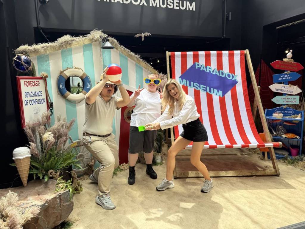 três pessoas posam com adereços de praia, incluindo uma bola de praia e uma pá, em frente a uma espreguiçadeira gigante às riscas e a uma cabana de praia azul no Paradox Museum London