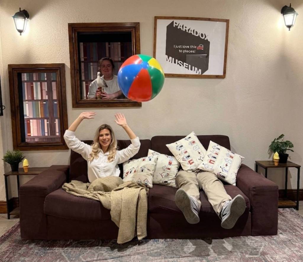 a woman throw a beach ball on the 'paradox sofa' where the illusions makes it look like the top half your body is one side and your legs come out the other side