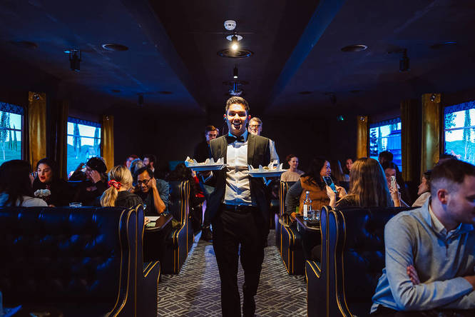 a waiter holding plates of food in a train carriage set at The Murder Express