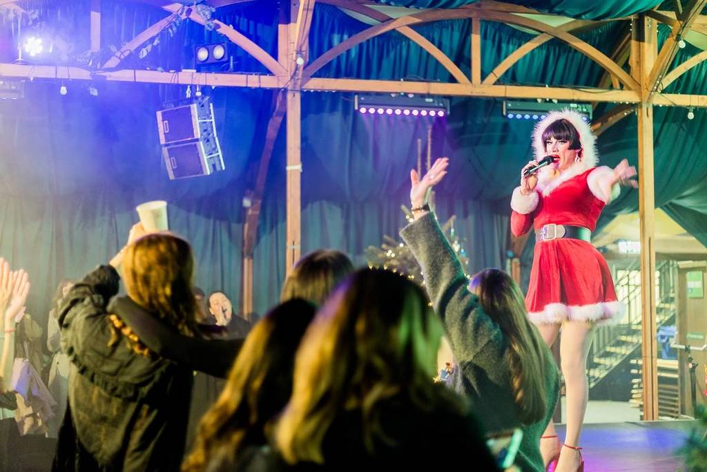 a drag star in a santa style outfit performs to a crowd with their hands in the air