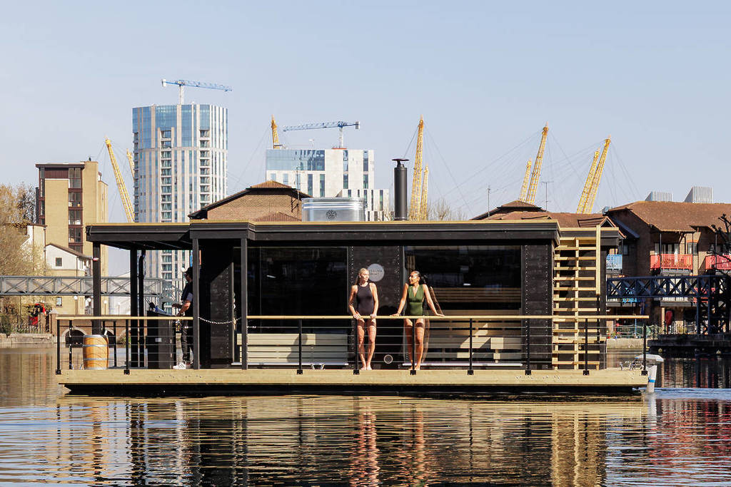 Zwei Frauen in Badekleidung stehen auf dem Balkon von Londons neuer schwimmender Sauna, mit den Docklands im Hintergrund