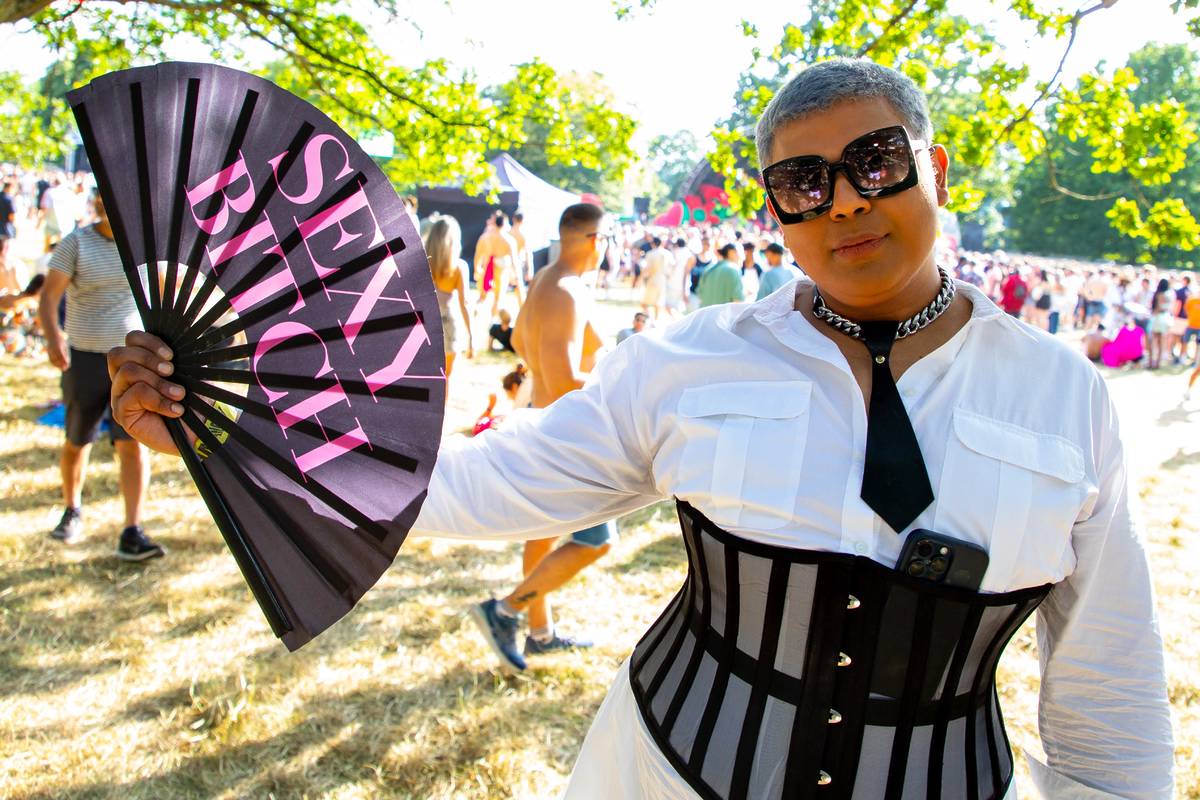 A stylish person poses confidently at As One In The Park Festival 2024. They wear a white button-up shirt with a black tie, a black-and-sheer corset, chunky chain necklace, and large black sunglasses. They hold an open black fan that reads “SEXY BITCH” in bold pink letters. A smartphone is tucked into the corset. In the background, a crowd enjoys the sunny event under trees and tents.