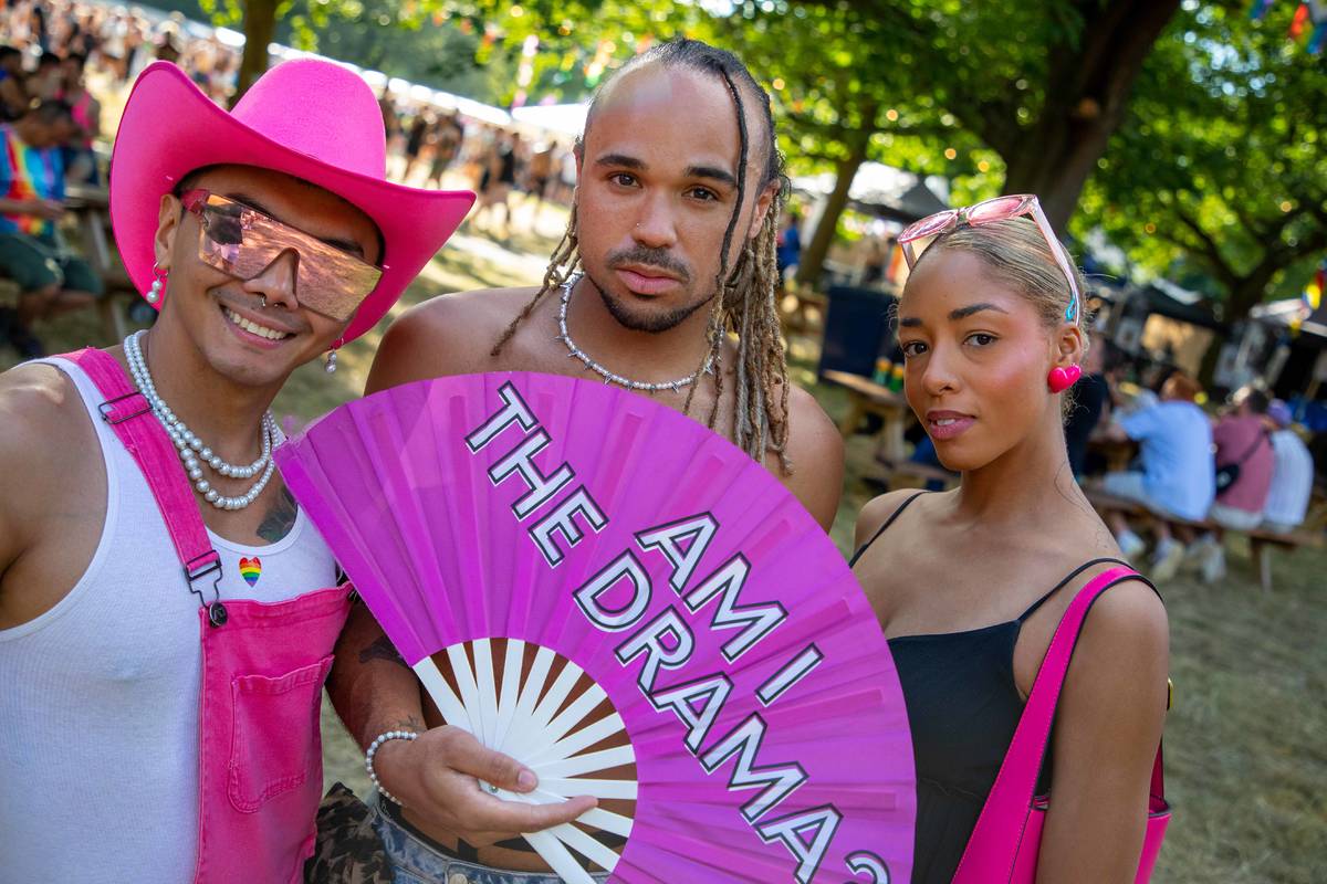 Three people pose at As One Festival 2024. One wears a pink cowboy hat and overalls, another holds a fan that says “AM I THE DRAMA?”, and the third wears a black dress with pink accessories. Trees and crowds fill the background.