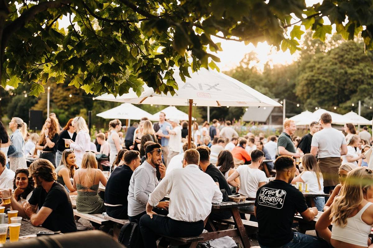 crowds of people sat and standing in a cafe seating area in a park with the setting sun casting a golden glow over everyone