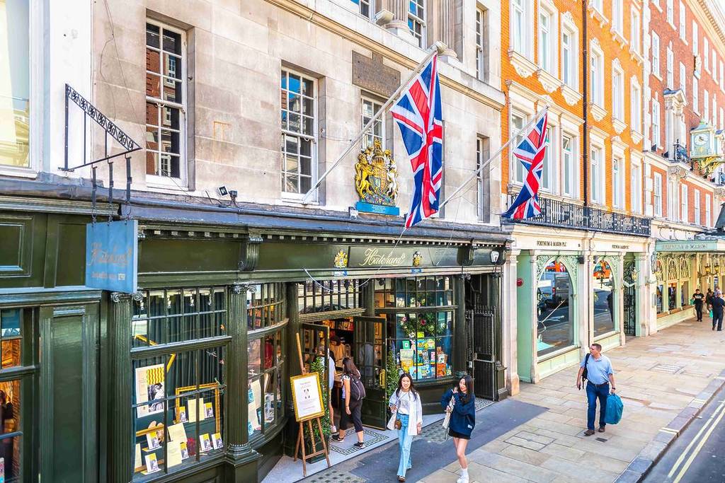 Exterior of Hatchards bookshop, taken from a slight angle. Sunny day, street level, people walking by. Shop front's splendent and decadent. UK flags hang next to a royal coat of arms.