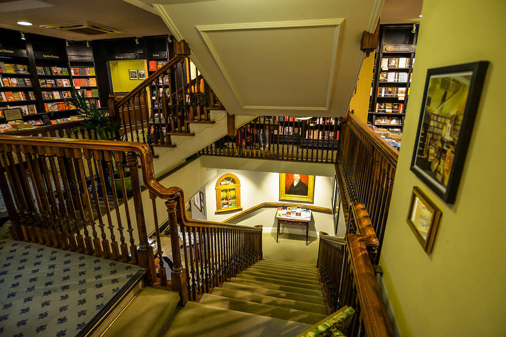 Interior of the bookshop. Yellow-tinged hue to the picture, evoking old vintage vibes. Decadent wooden banisters curl and cut across each other, levels of stairs all over.
