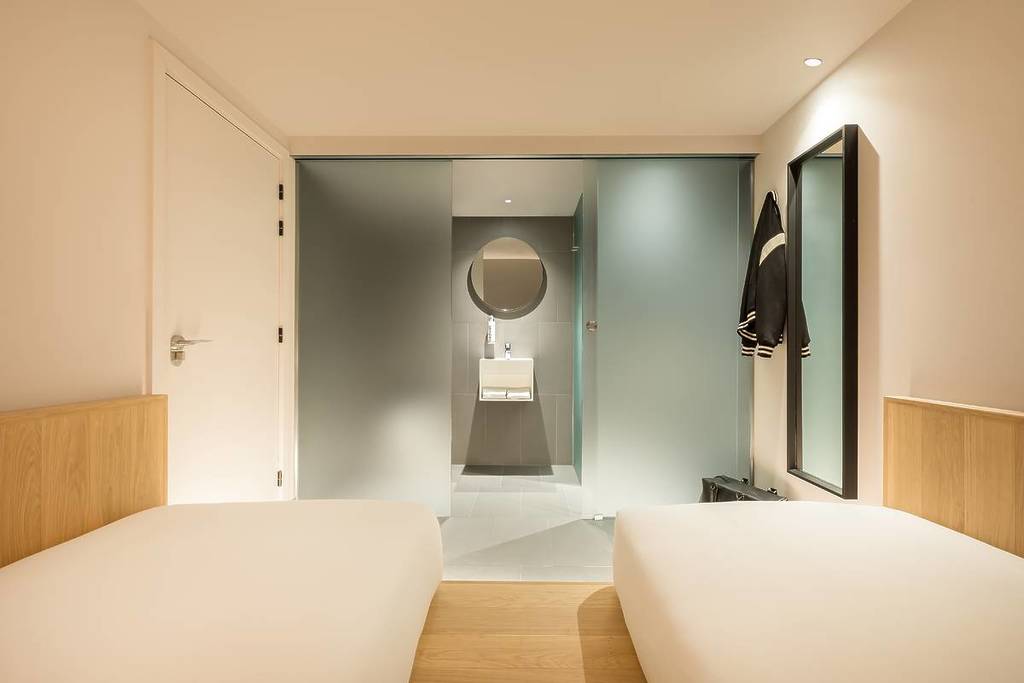 A light, airy yet small hotel room. Clean white plain walls. Two twin beds, from where the photos' taken, look down said beds, facing a bathroom: frosted glass dividers, round mirrors, and ultra modern lighting.